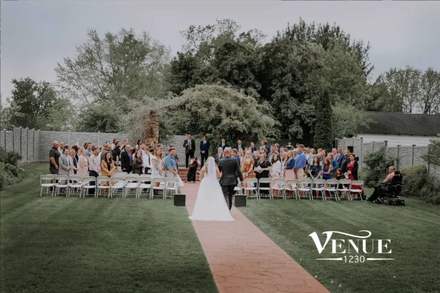 Outdoor wedding ceremony with guests seated along a pink aisle, bride in white gown approaching groom amid greenery and stone walls.